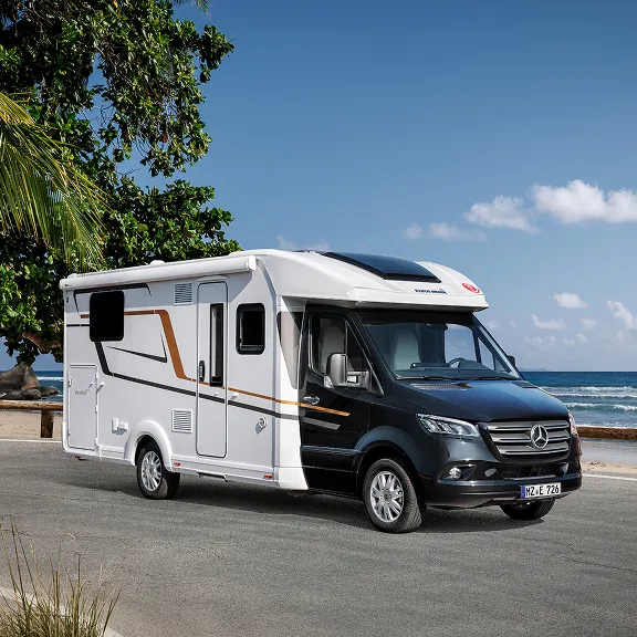 Weißes Wohnmobil an Küstenstraße mit Palmen, im Hintergrund Strand und Meer bei blauem Himmel.