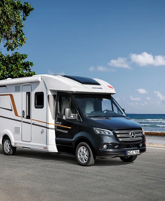 Teilintegriertes Mercedes-Wohnmobil auf einer Küstenstraße direkt am Meer unter Palmen.