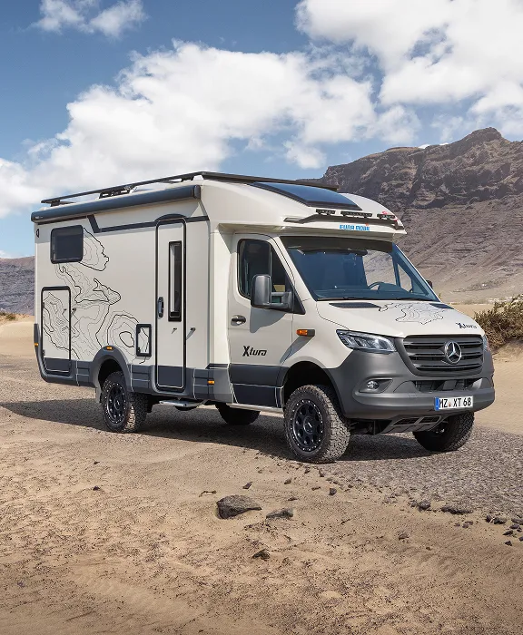 Geländegängiges Eura Mobil XTura Wohnmobil auf einem Sandweg vor einer bergigen Küstenlandschaft.